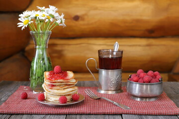 Fototapeta na wymiar On a wooden table there is a stack of pancakes with fresh raspberries and jam near a bowl of ripe berries, a glass of tea and a vase of flowers.