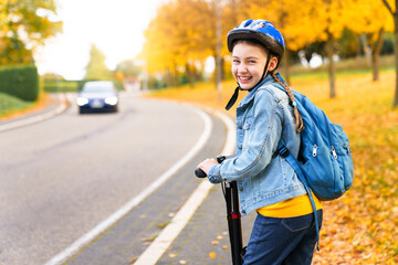 Road to school. Happy kid riding scooter on autumn city street. Child girl with protective helmet and blue backpack on safety bikeway. Back to school concept. Ecological transportation and lifestyle - obrazy, fototapety, plakaty
