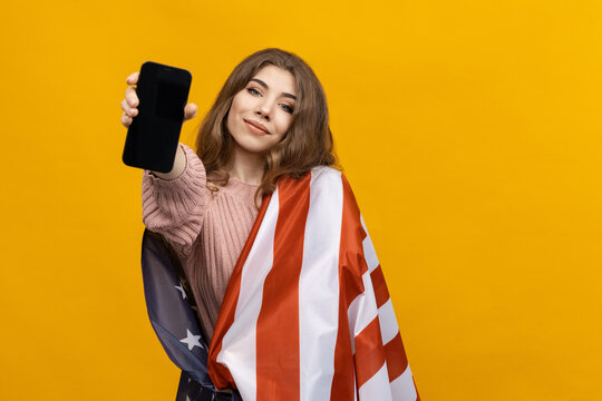 A young woman with an American flag on her shoulders holds a mobile phone on a yellow background. The ability to place ads on a smartphone. Mockup
