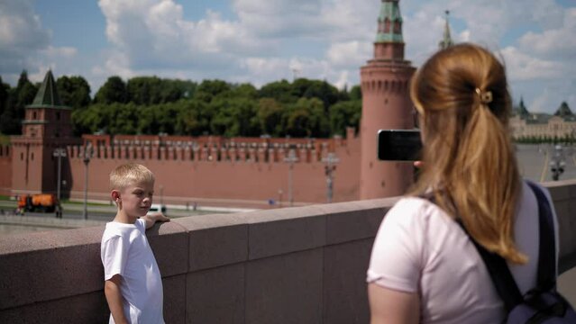 A mother takes pictures of her son on her phone on the bridge against the background of the Kremlin and Red Square. Tourists walk around the city and take pictures.