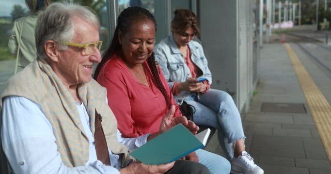 Multiracial senior people sitting and waiting together at tram station in the city - Senior man reading a book