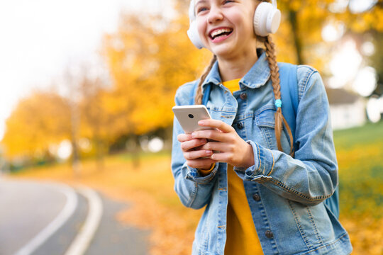 Happy school child using smartphone on autumn town street. Teenager in headphones listening to music outdoors. Girl smiling, having fun. Kid messaging online. Technology lifestyle, communication