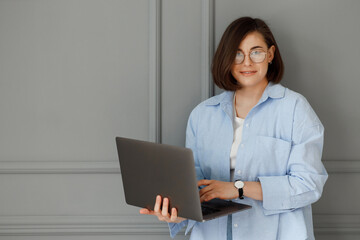 Beautiful young businesswoman with glasses on her eyes is wearing a white t-shirt and a blue shirt is holding her laptop in her hands on a grey blackground