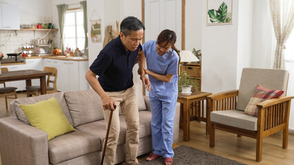 korean woman in-home care attendant assisting senior male to stand up from the sofa at home. the senior patient using a crutch is feeling great pain in his knee joints