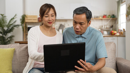 asian smiling daughter and confused elderly father sitting together with a laptop computer on living room sofa at home. she teaches him how to surf the internet with gestures