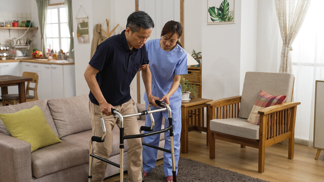 Asian Japanese Senior Stroke Patient Undergoing Rehab Exercise With A Walker At Home. The Woman Nursing Aide Assists Him During Home Visit