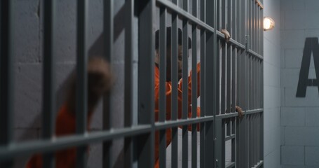 African American prisoner in orange uniform leans on jail cell bars. Neighbor criminal sticks out...