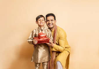 Happy young indian father and son wearing traditional kurta outfit holding red gift boxes celebrating of diwali festival isolated on beige background. Parent with child boy holding present.