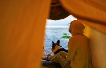 Traveler woman and her pet sitting inside tent and admiring nature view at tranquil beach. Experience the joy of exploring the world with your furry friend