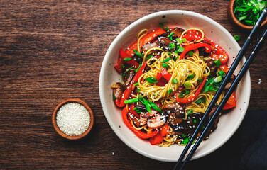 Stir fry noodles with vegetables: paprika, champignons, chives and sesame seeds in ceramic bowl. Wooden table background, top view