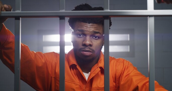 African American man in orange uniform looks at camera and kneads neck in prison cell. Prisoner serves imprisonment term in jail. Criminal in correctional facility or detention center. Portrait view.
