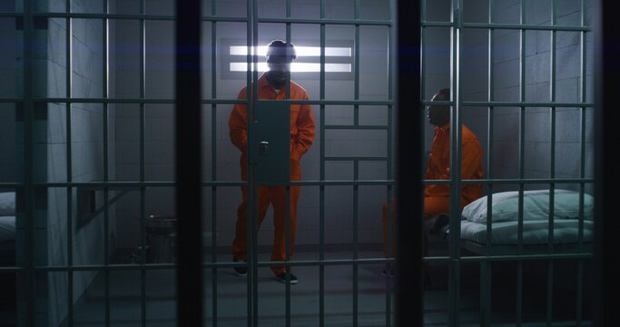 Two African American prisoners in orange uniforms talk. One man sits on bed, another walks in prison cell. Men serve imprisonment terms for crimes in jail, detention center or correctional facility.