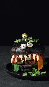 Monster burger with black bun, vegan patty, cheese, olive eyes and smoke on the black background. Funny Halloween food