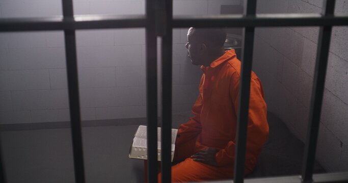 African American prisoner in orange uniform sits on bed behind bars, reads Bible in prison cell. Male criminal serves imprisonment term for crime in jail. Detention center or correctional facility.