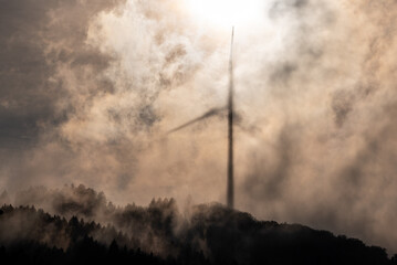 Windrad im Gegenlicht auf einem bewaldeten Hügel in einer Wolke, in die die Sonne den Schatten des...
