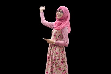 Close up portrait of smiling young Muslim woman wearing pink hijab with flower motif dress waving right arm up gesture and left hand holding phone. Happy and cheerful expression.