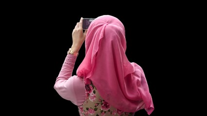Close up portrait of young Muslim woman back of head wearing pink hijab and dress with red rose motif taking picture and video with phone camera.