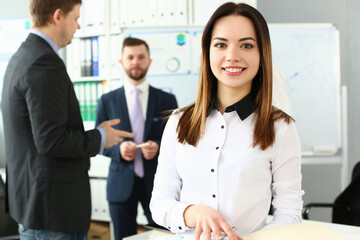 Portrait of smiling confident business woman standing in office with colleagues in background. Student successful happy entrepreneur employee looking at camera