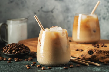 Ice coffee in a tall glass with cream poured over, ice cubes and beans on a dark concrete table.