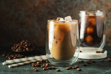 Ice coffee in a tall glass with cream poured over, ice cubes and beans on a dark concrete table.