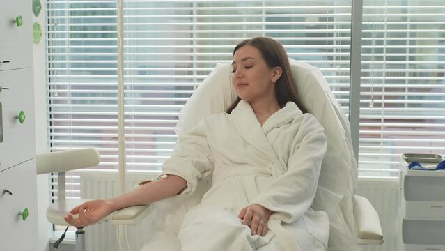 Tracking shot portrait of pretty young woman patient in white bathrobe with IV catheter in vein receiving vitamin treatment in beauty salon lying on comfy medical armchair. Shooting in slow motion.