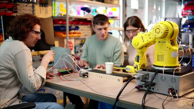 Young happy engineers programming an yellow robotic arm in the workshop to grab cardboard water glass, computer programming training for coffee preparation, celebrating success