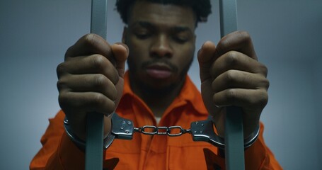 African American prisoner in orange uniform keeps hands in handcuffs on jail cell bars. Guilty...