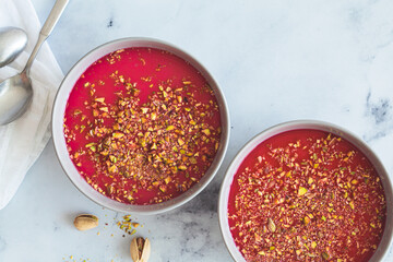 Malabi traditional Arabic dessert. Milk pudding with pink syrup and pistachio in gray bowl, white marble background.
