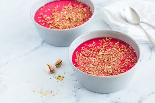 Malabi traditional Arabic dessert. Milk pudding with pink syrup and pistachio in gray bowl, white marble background.