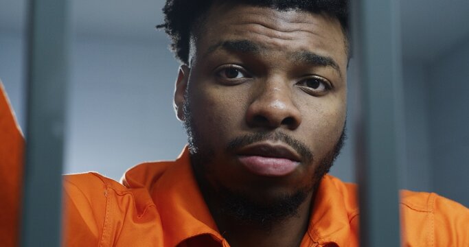 African American man in orange uniform stands behind prison cell bars and looks at camera. Prisoner serves imprisonment term in jail. Criminal in correctional facility or detention center. Portrait.