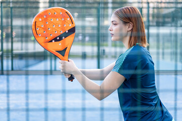 Photo of a girl behind a fence of an outdoor paddle tennis court that is posing with a racket to receive a serve. Concept of women playing paddle. Paddle for women.