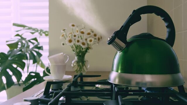 A green teapot boiling on a gas stove over an open flame. Kettle with boiling water on a gas stove with the steam plume clearly visible. Teakettle boiling with steam emitted from the spout. Sunlight f