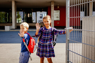 Back to school. Happy siblings with backpacks, primary school kids, waving their hands on their way...