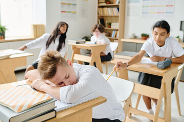 Schoolboy sleeping at desk at lesson while his classmate waking him up