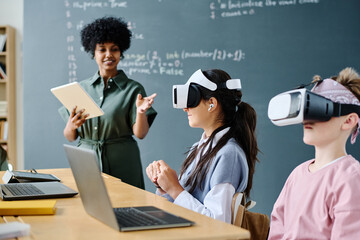 Students using VR glasses while sitting at table and listening to teacher at IT lesson