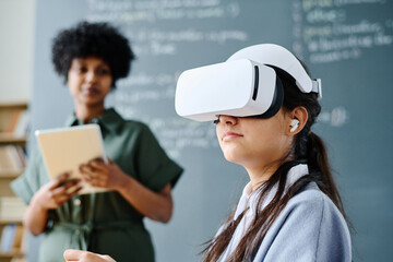 Student in VR glasses sitting at lesson with teacher explaining material in background