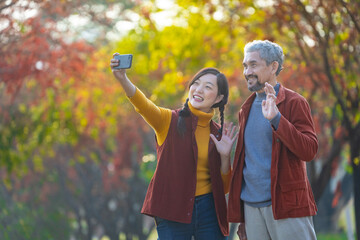 Happy Asian daughter and her senior father are walking together in public park during autumn with maple and ginkgo tree while taking selfie for fall color travel destination and family happiness