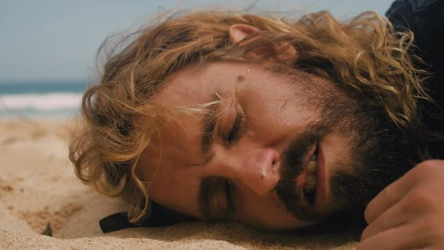 Male person lying on sand beach suffering from hangover. Homeless man in depression listening to wave. Peaceful noise of ocean. Sun stroke. Tired smile of solo traveler. Stray tramp sleeping outdoors.