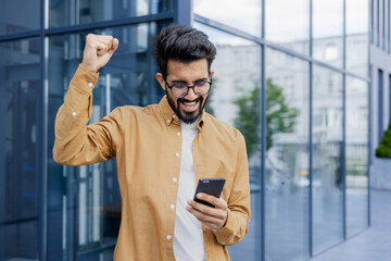 Young successful hispanic man in casual clothes outside office building received online win...