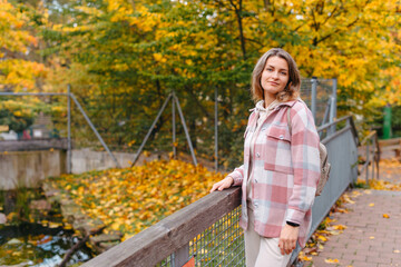 Portrait Of Cute Young Woman In Casual Wear In Autumn, Standing On Bridge Against Background Of An Autumn Park And River. Pretty Female Walking In Park In Golden Fall. Copy Space. Smiling Girl In The