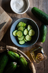 Vista desde arriba de un bol con rodajas de pepino, junto a una cesta con pepinos, una tabla de cortar, un bol con sal y una aceitera de vidrio