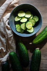 Vista cenital de un bol con rodajas de pepino y unos pepinos enteros sobre una mesa de madera