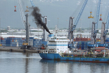 Black smoke as air pollution coming out from funnel of tanker vessel leaving the port. Behind is container terminal with boxes and mobile cranes.