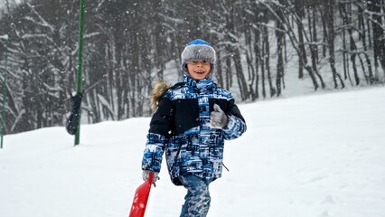 Fototapeta na wymiar Joyful boy running up snowy hill with sleds in hand, ready to enjoy the winter fun of sledding down