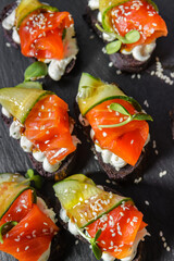 Canapes of black bread, red fish and cucumbers, seasoned with sauce and sprinkled with sesame seeds, lie on a tray. A fragment of the serving.