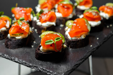 Canapes of black bread, red fish and cucumbers, seasoned with sauce and sprinkled with sesame seeds, lie on a tray. A fragment of the serving.