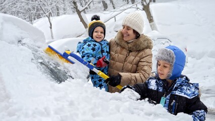 Smiling mother and her baby son work together to remove snow from their car with a brush after a blizzard. The concept of daily routine and vehicle maintenance