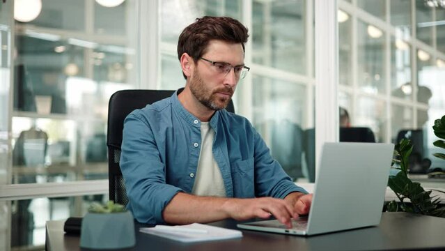 Male manager handsome man, office worker manager, wearing blue shirt, corrects glasses working on laptop. It programmer working on project, remote work, noting down important details on paper.
