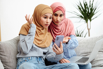 Two Arab women are having trouble communicating with the banking system. Sit together on the couch and look at the phone. Girls have expressions of surprise and shock on their faces. Throw up hands.
