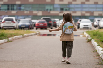 A little girl with a backpack behind her back is walking along a path with puddles to a building through a parking lot of cars. Copy space.